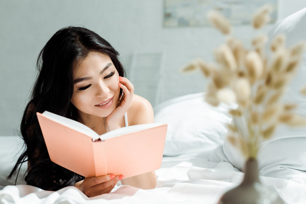 enfoque selectivo de positiva chica asiática lectura libro mientras está acostado en la cama en casa
  - Foto, Imagen