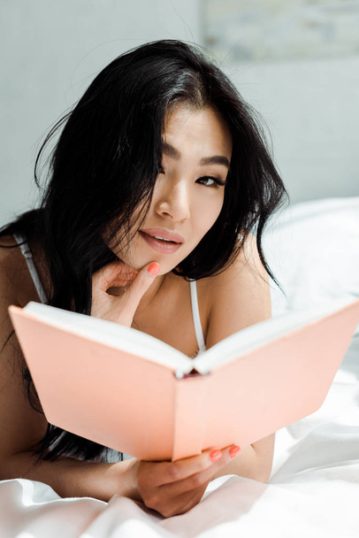 selective focus of pretty thai woman holding book and looking at camera  - Foto, Imagen