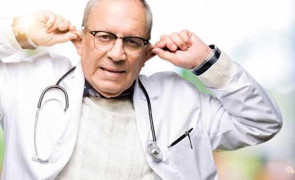 Hombre guapo médico senior con abrigo médico Sonriendo tirando de las orejas con los dedos, gesto divertido. Problema de audición
 - Foto, imagen