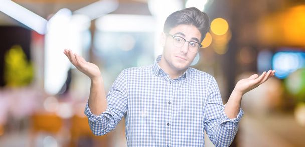 Young handsome man wearing glasses over isolated background clueless and confused expression with arms and hands raised. Doubt concept. - Photo, Image