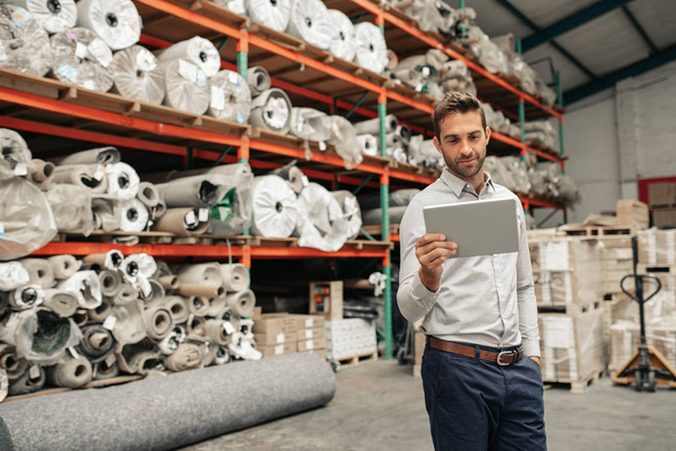 Smiling manager using a digital tablet while standing on the floor of a warehouse with stacks of carpets and textiles on shelves in the background - Photo, Image