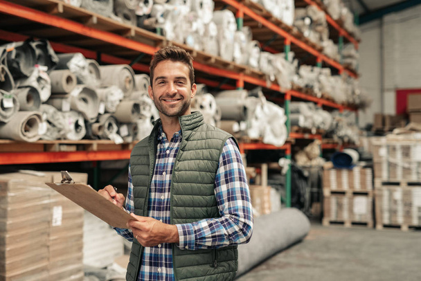Portrait of a smiling carpet warehouse worker checking inventory with a clipboard with stacks of stock on shelves in the background - Photo, Image