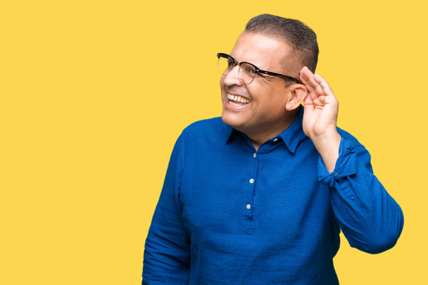 Arabischer Mann mittleren Alters mit Brille vor isoliertem Hintergrund, der lächelnd mit der Hand über dem Ohr Gerüchten oder Klatsch lauscht. Konzept für Gehörlosigkeit. - Foto, Bild