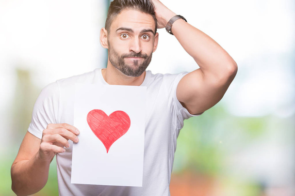Handsome young man holding card with red heart over isolated background stressed with hand on head, shocked with shame and surprise face, angry and frustrated. Fear and upset for mistake. - Фото, изображение