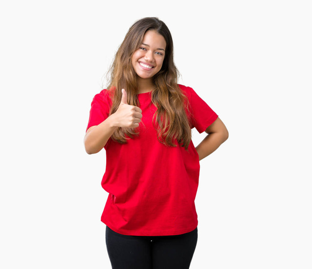 Jonge mooie brunette vrouw met rode t-shirt over geïsoleerde achtergrond gelukkig duimschroef opwaarts gebaar met de hand te doen. Goedkeuring van de expressie te kijken naar de camera toont succes. - Foto, afbeelding