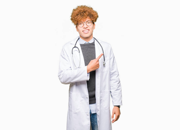 junger, gutaussehender Arzt in medizinischem Mantel, fröhlich mit einem Lächeln im Gesicht, das mit der Hand und dem Finger zur Seite zeigt, mit glücklichem und natürlichem Gesichtsausdruck - Foto, Bild
