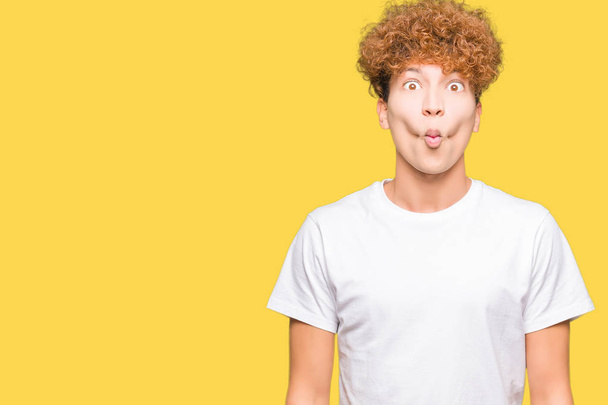 Young handsome man with afro hair wearing casual white t-shirt making fish face with lips, crazy and comical gesture. Funny expression. - Photo, Image