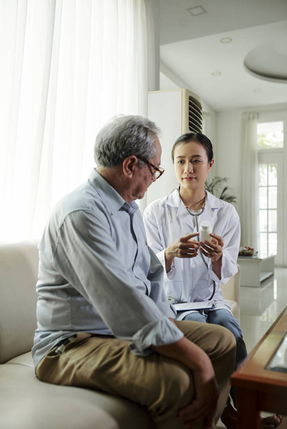 Mladý asijský lékař sedící na pohovce s vyšším mužem, který navrhuje dobré pilulky před nemocí a mluví o jeho léčbě při návštěvě doma - Fotografie, Obrázek