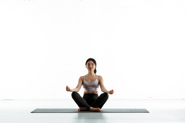 calm asian woman sitting on yoga mat and doing exercise on white - Фото, зображення