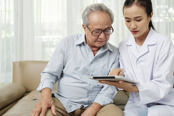 Médica asiática mostrando tratamento on-line em comprimido digital para paciente sênior enquanto eles sentados juntos no sofá
 - Foto, Imagem