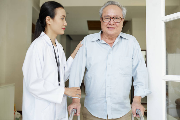 alter Mann mit Brille, der mit Rollator steht und in die Kamera schaut, während eine Krankenschwester in weißem Mantel ihm hilft, sich zu bewegen - Foto, Bild