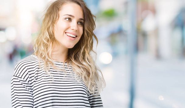 Mooie jonge blonde vrouw draagt strepen trui over geïsoleerde achtergrond op zoek weg aan kant met glimlach op het gezicht, natuurlijke wijze van uitdrukking. Lachen vertrouwen. - Foto, afbeelding