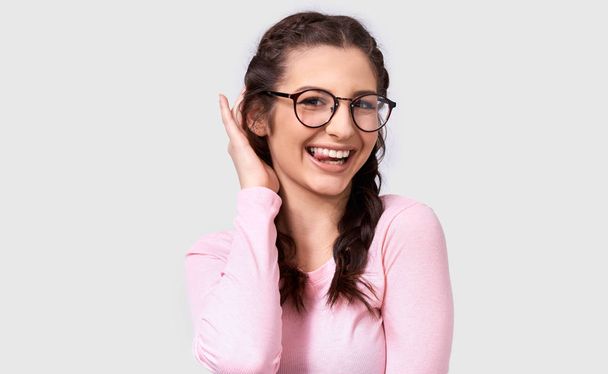 Retrato de una joven divertida mostrando su lengua, parpadeando con un ojo y llevando gafas redondas transparentes de moda mientras posaba en la pared blanca del estudio. Hembra sonriente positiva haciendo mueca. Gente
 - Foto, imagen