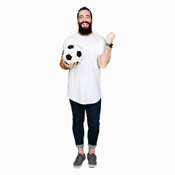 Νεαρό άνδρα με μακριά μαλλιά και αρκούδα κρατώντας την μπάλα ποδοσφαίρου ποδοσφαίρου ουρλιάζοντας περήφανος και γιορτάζει τη νίκη και την επιτυχία πολύ ενθουσιασμένος, ζητωκραυγάζει συγκίνηση - Φωτογραφία, εικόνα