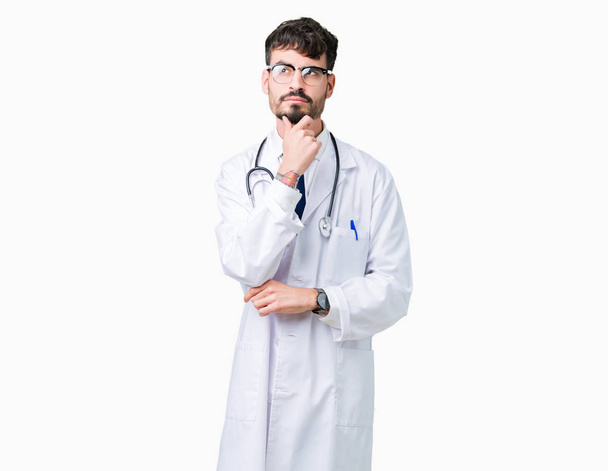若い医者あごを考えて質問、物思いにふける式の手で隔離された背景に病院のコートを着た男。思いやりのある顔をして笑っています。疑いのコンセプト. - 写真・画像