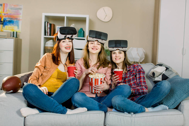 Három fiatal vonzó nő visel fülhallgatót VR virtuális valóság látás szemüveg, miután fél otthon, evés pattogatott kukorica - Fotó, kép