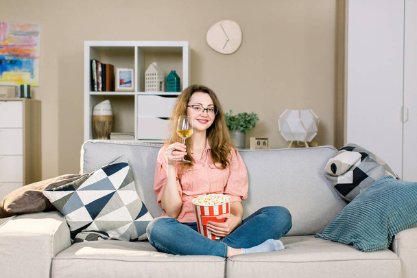Молода усміхнена жінка вдома сидить на дивані і дивиться телевізор, вона тримає келих з вином і їсть попкорн
 - Фото, зображення