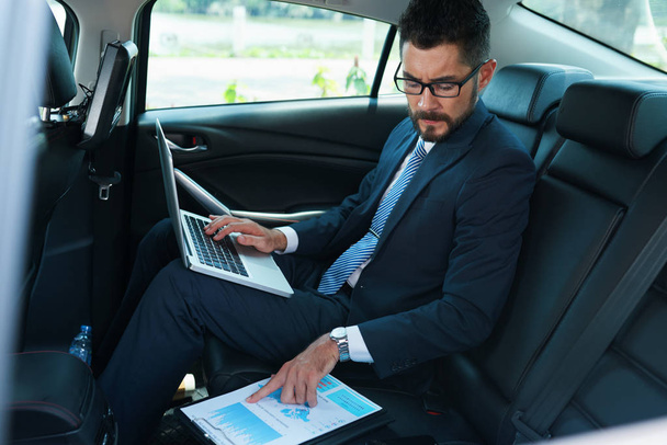 Serieuze financiële manager zittend op achterbank van de auto en werken met documenten - Foto, afbeelding
