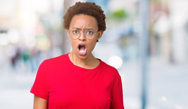 Όμορφη νεαρή αφρικανική αμερικανική γυναίκα φοράει γυαλιά, πέρα από το απομονωμένο υπόβαθρο στο πρόσωπό του σοκ, αναζητούν σκεπτικός και σαρκαστική, εξέπληξε με ανοιχτό το στόμα - Φωτογραφία, εικόνα