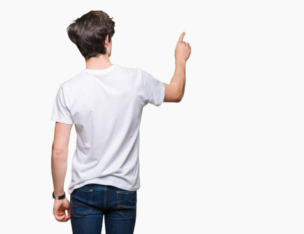 Joven hombre guapo con camiseta blanca casual sobre fondo aislado posando hacia atrás apuntando hacia atrás con la mano del dedo
 - Foto, Imagen