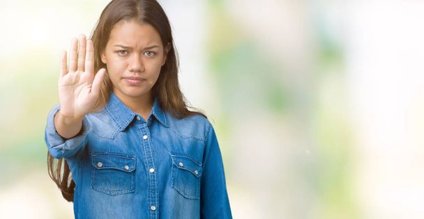Young beautiful brunette woman wearing blue denim shirt over isolated background doing stop sing with palm of the hand. Warning expression with negative and serious gesture on the face. - Photo, Image