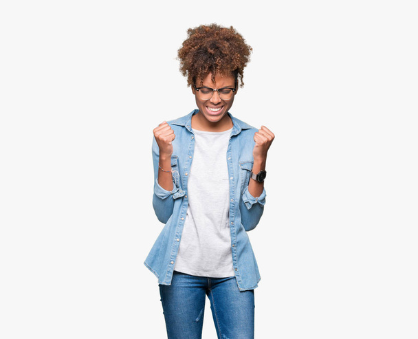 Hermosa mujer afroamericana joven con gafas sobre fondo aislado muy feliz y emocionado haciendo gesto ganador con los brazos levantados, sonriendo y gritando por el éxito. Concepto de celebración
. - Foto, imagen