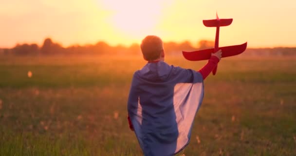 De jongen presenteert zichzelf als piloot en loopt in het veld bij zonsondergang met een vliegtuig - Video