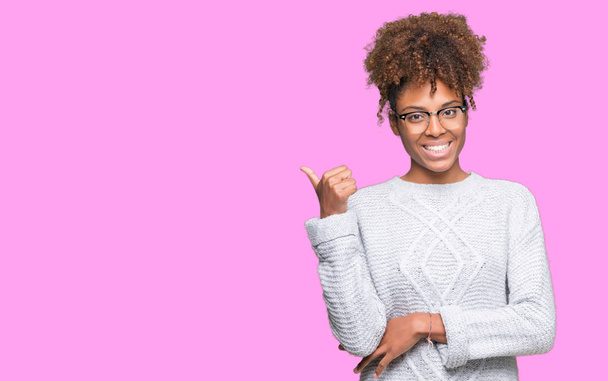 Красивая молодая африканская американка в зимнем свитере на изолированном фоне улыбается со счастливым лицом, глядя и указывая в сторону с большим пальцем вверх
. - Фото, изображение