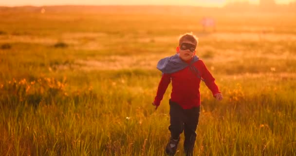 Ein Junge in Anzug und Superheldenmaske läuft bei Sonnenuntergang über das Feld auf dem Rasen - Filmmaterial, Video