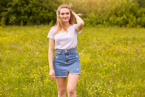Outdoor shot of pretty blond woman wearing white casual t shirt and denim skirt, posing in meadow, touching her fair hair, looking at camera with pleasant facial exprassion. Happyness concept. - Fotoğraf, Görsel