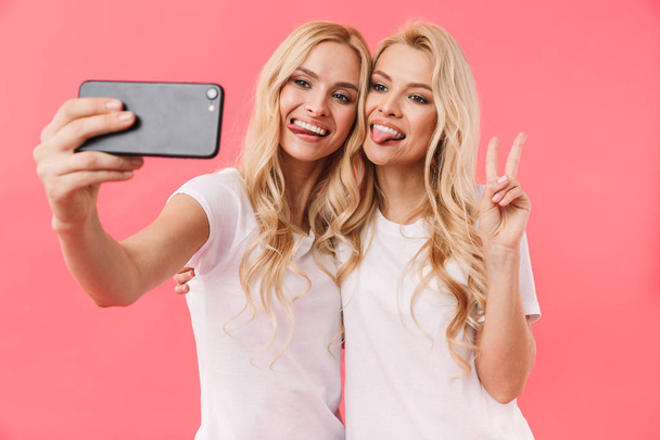 Happy blonde twins wearing in t-shirts making selfie on smartphone - Фото, изображение