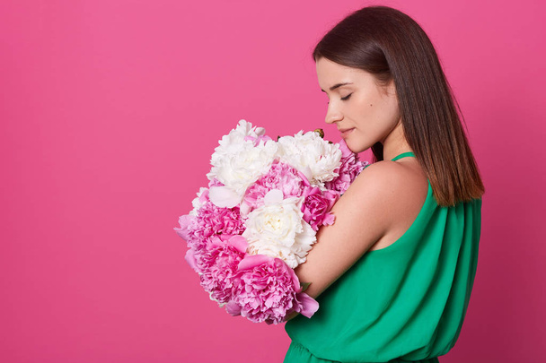 Indoor studio shot of sweet adorable young woman standing isolated over pink background with closed eyes, having peaceful look, holding white and pink flowers in hands. Copyspace for advertisement. - Fotografie, Obrázek