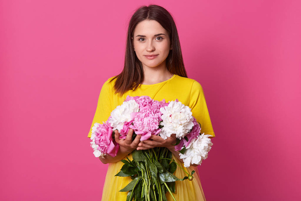 Portrait of tender magnetic black haired lady wearing bright yellow items of clothes, looking directly at camera, having good mood, holding bouquet of white and pink peonies. Spring concept. - Фото, изображение