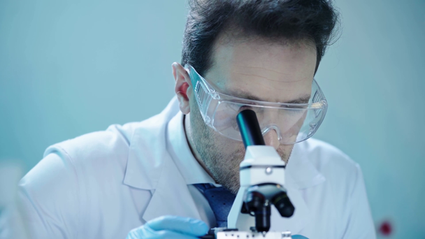 όμορφος επιστήμονας σε προστατευτικά γυαλιά κάνοντας ανάλυση με μικροσκόπιο και γράφοντας τα αποτελέσματα στο εργαστήριο - Πλάνα, βίντεο