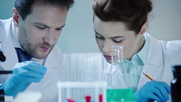 scientist making analysis in laboratory and talking to pretty doctor sitting near and writing down results - Filmagem, Vídeo