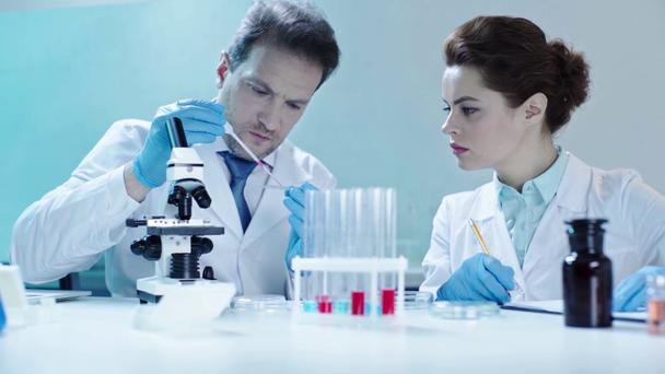 handsome scientist taking red liquid from test tube while talking to pretty doctor writing results - Footage, Video