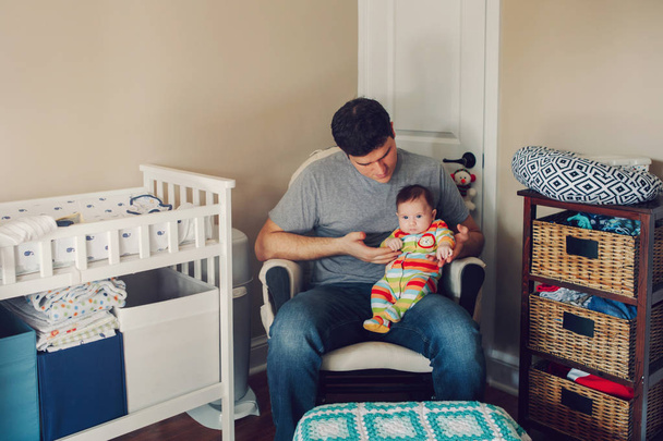 Beyaz bir baba, yeni doğmuş bir bebekle çocuk odasında oturuyor. Erkek bir ebeveyn çocuğunu kucağında tutuyor. Otantik yaşam tarzı çok sevimli bir an. Bekar baba aile hayatı konsepti.  - Fotoğraf, Görsel