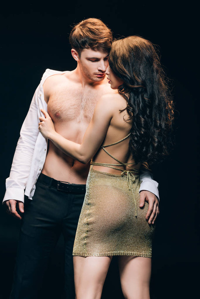 sexy brunette woman undressing boyfriend with muscular torso isolated on black - Valokuva, kuva