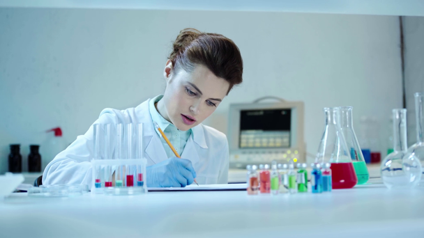 attentive scientist looking at blue liquid in test tube and writing down results in laboratory - Footage, Video