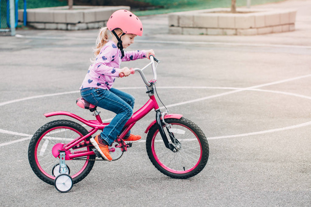 Porträt eines lächelnden kaukasischen Vorschulmädchens, das an einem Frühlingssommertag auf einem rosafarbenen Fahrrad mit Helm in der Hinterhofstraße unterwegs ist. Saisonale Kinderaktivitätskonzepte. Gesunder Lebensstil in der Kindheit - Foto, Bild