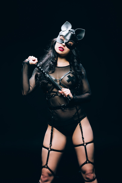 passionate hot brunette woman in bdsm costume and mask with flogging whip isolated on black - Foto, imagen