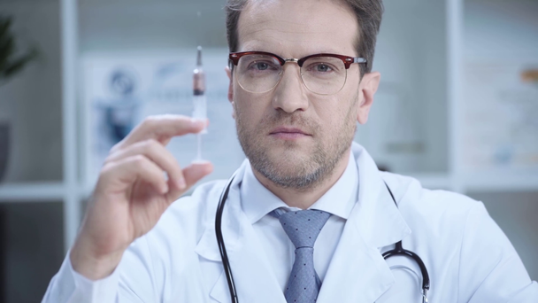 pohledný doktor ve skleničkách zobrazující injekční stříkačku s tekutinou a s pohledem na fotoaparát - Záběry, video