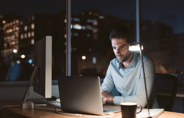 Young businessman working at his desk in an office late at night with city lights glowing in the background - Photo, image
