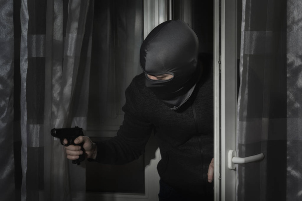 Κλέφτης με ένα όπλο στο χέρι του και με ένα καπέλο με μια σχισμή για τα μάτια τρυπώνει μέσα στο σπίτι μέσα από την πόρτα τη νύχτα στο φεγγαρόφωτο - Φωτογραφία, εικόνα