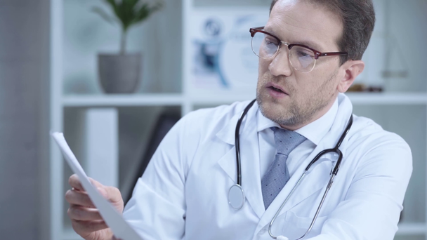 serious doctor with stethoscope talking while looking at paper with cardiogram - Felvétel, videó