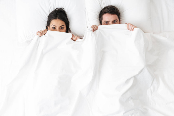Young loving couple lies in bed hide covering under blanket. - Фото, изображение