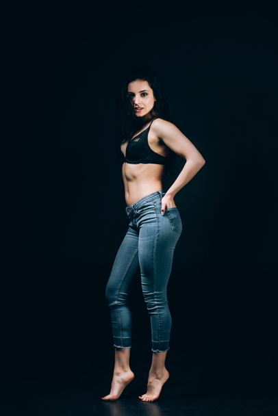 sexy brunette barefoot woman in blue jeans and bra posing with hands in pockets isolated on black - Zdjęcie, obraz