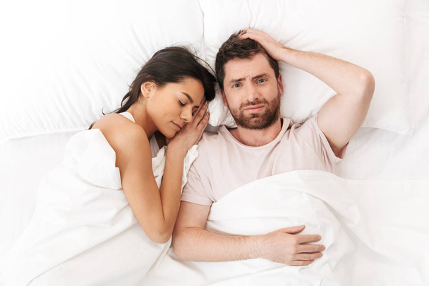 Displeased confused young man lies in bed under blanket near sleeping woman. - Foto, Bild