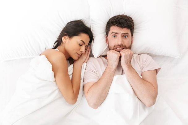 Mécontent jeune homme confus se trouve dans le lit sous la couverture près de la femme endormie
. - Photo, image