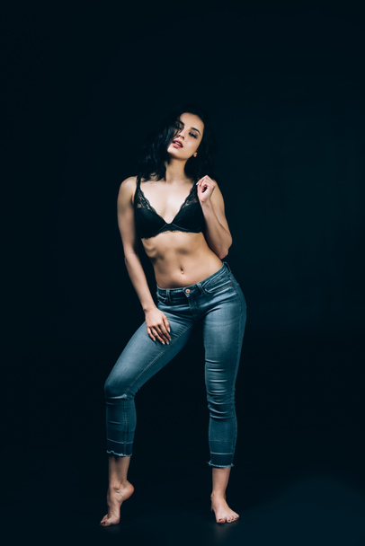 sexy brunette barefoot woman in blue jeans and bra posing isolated on black - Photo, image